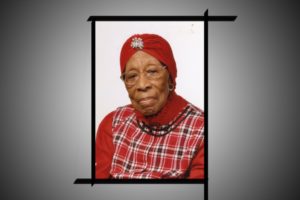 Still photograph of Grandmother Taylor, wearing a red plaid tunic, red turtleneck, and red turban with a jewel in the middle